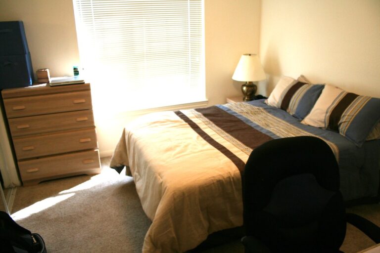 Cosy bedroom with a made bed, bedside lamp and soft sunlight through window blinds