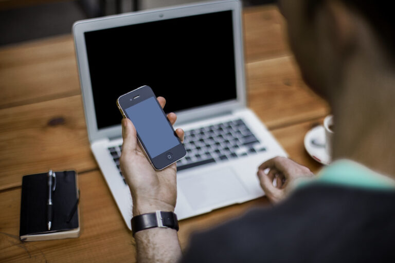 Person holding a smartphone at a wooden desk beside an open laptop and notebook