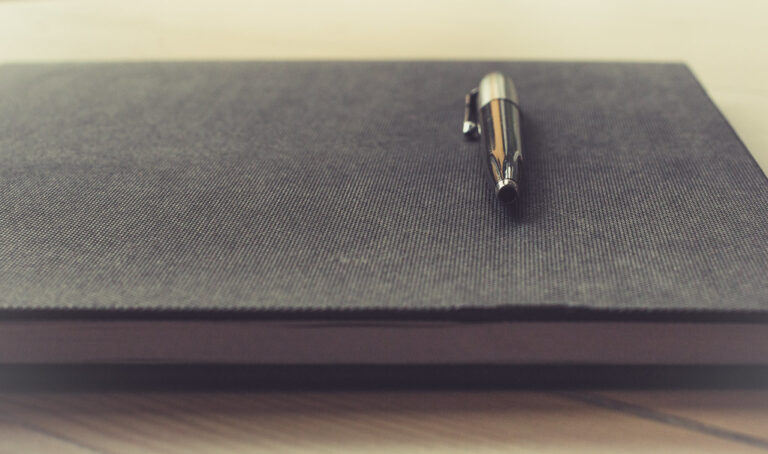 Closed dark notebook with a silver pen resting on a light wooden desk