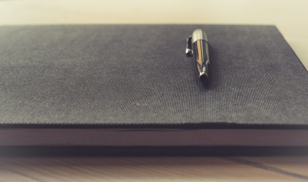 Closed dark notebook with a silver pen resting on a light wooden desk