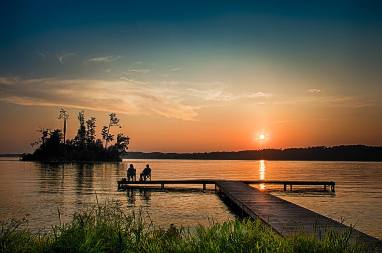 Two people sitting on a wooden dock by a calm lake at a golden sunset
