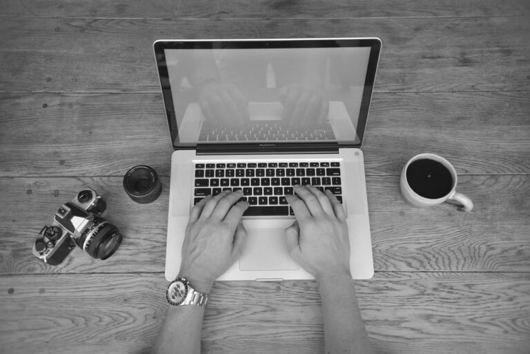 Black and white photo of hands typing on a laptop beside a coffee cup