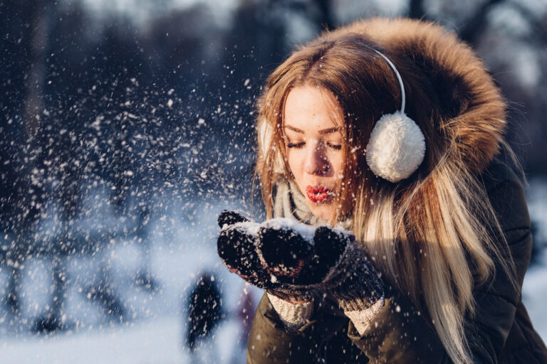Young woman in earmuffs and a warm hood blowing snow from her mittens