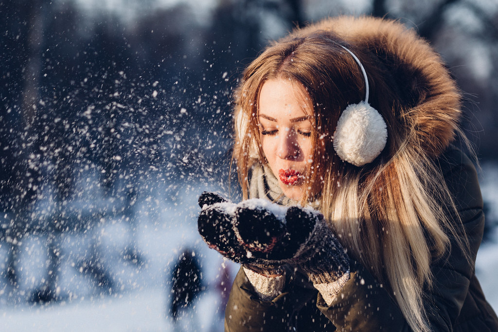 Young woman in earmuffs and a warm hood blowing snow from her mittens