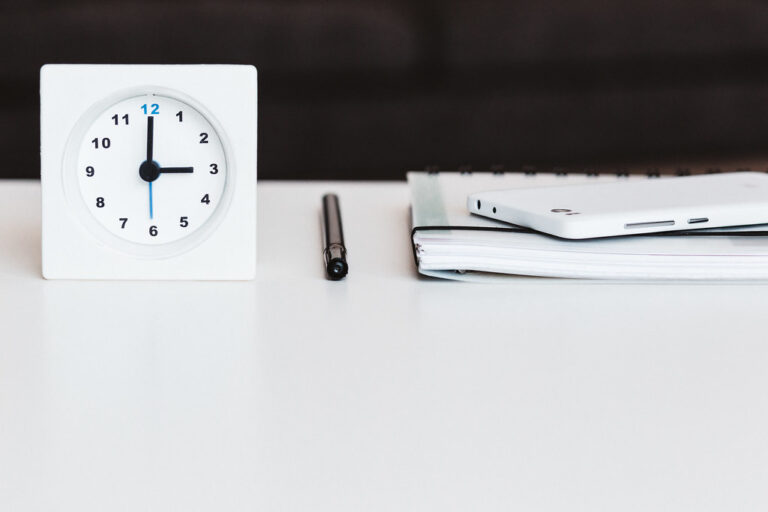 White desk clock, black pen and notebook arranged neatly on a white desk