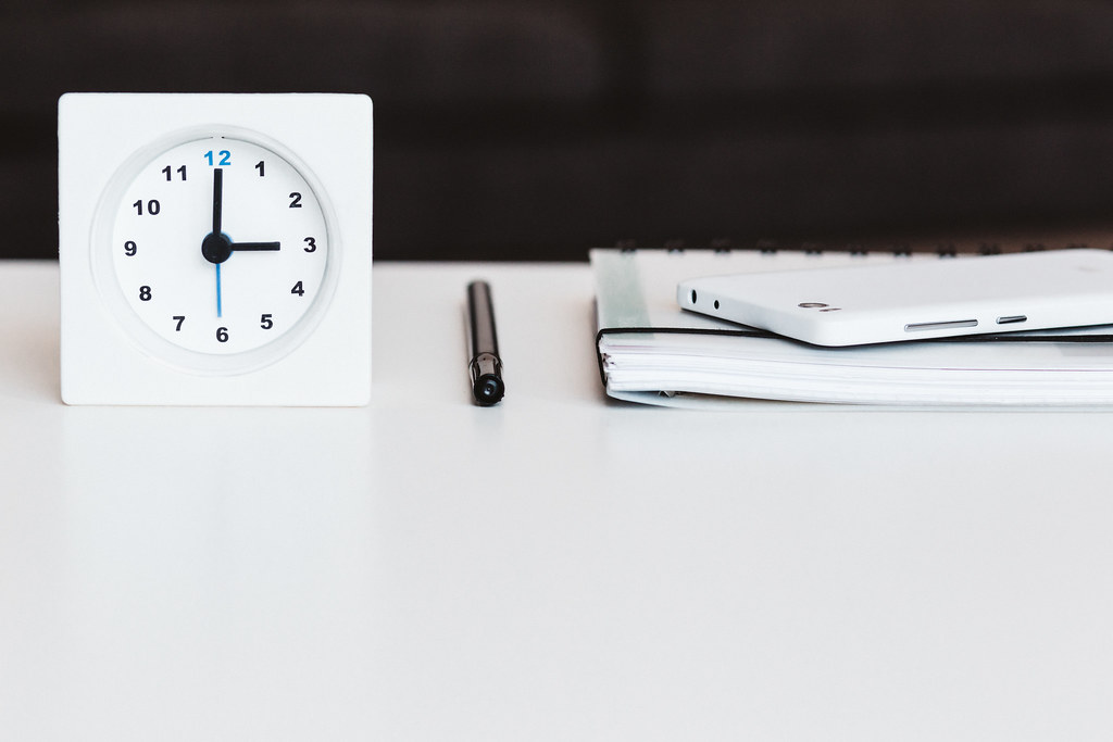 White desk clock, black pen and notebook arranged neatly on a white desk