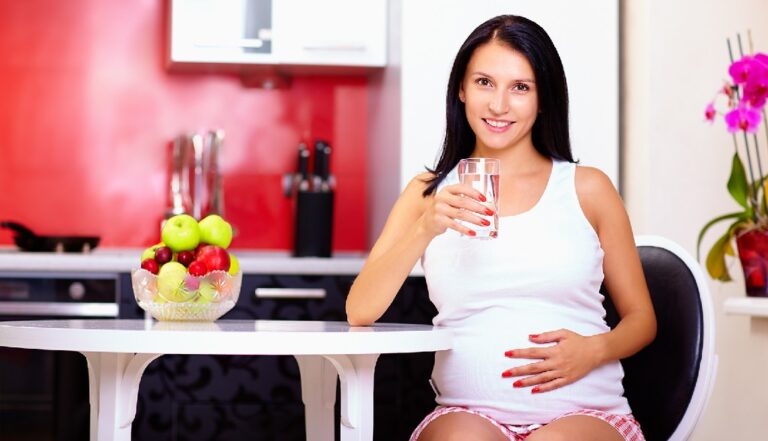 Smiling woman holding a glass of water at a kitchen table with fresh apples