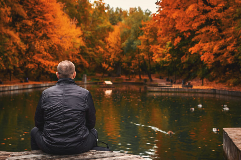 Man sitting quietly by a calm park pond surrounded by bright autumn trees