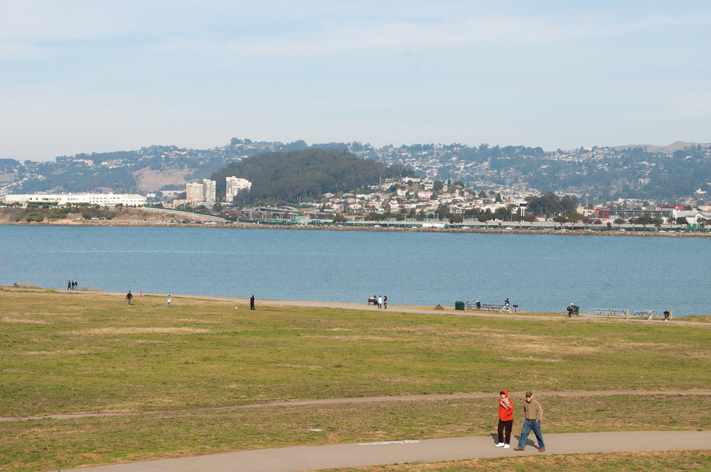 Two people walking on a paved park path beside a calm blue bay