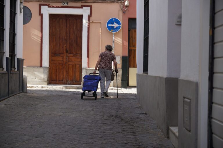 Older woman with a walking cane pulling a blue shopping cart down a quiet street