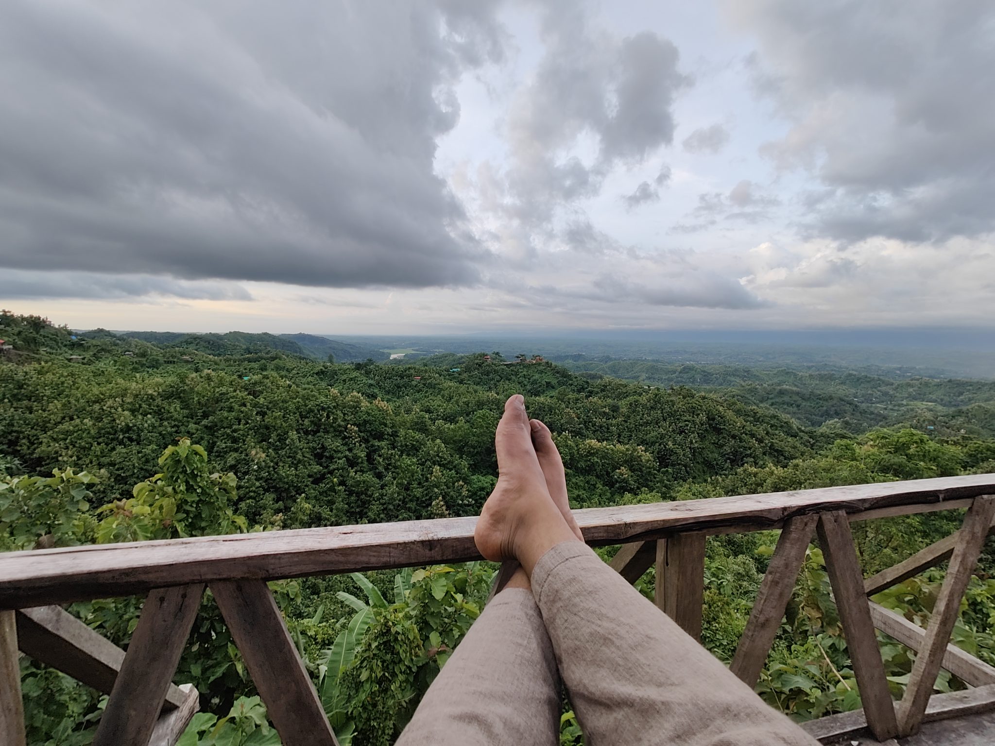Bare feet resting on a wooden railing above green forested hills under cloudy sky