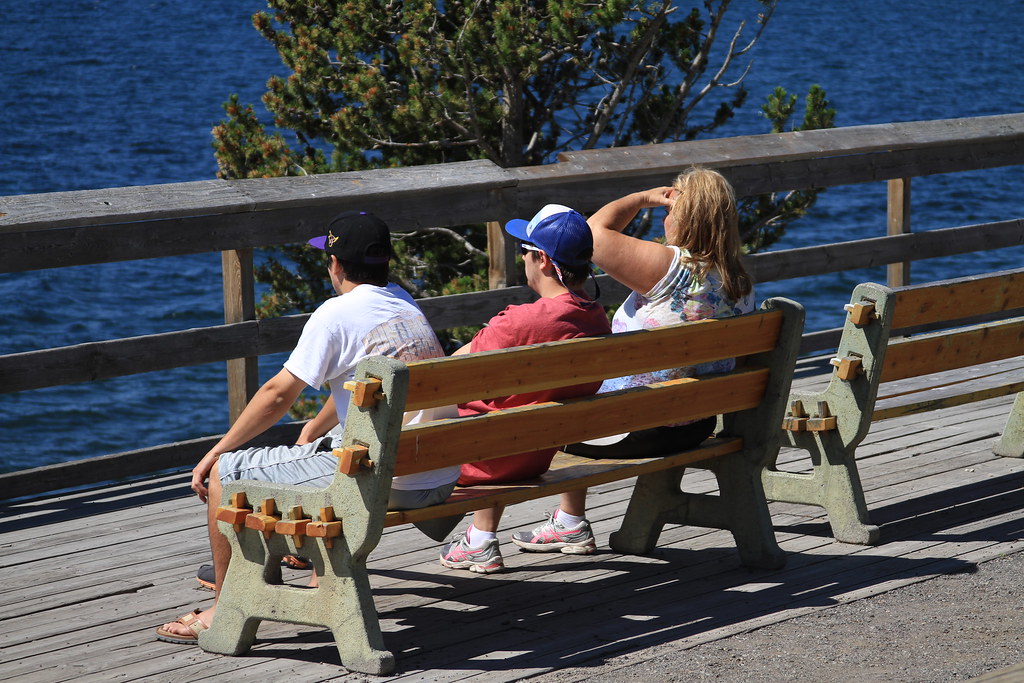 Three visitors resting on a wooden bench on a sunny deck beside a blue lake